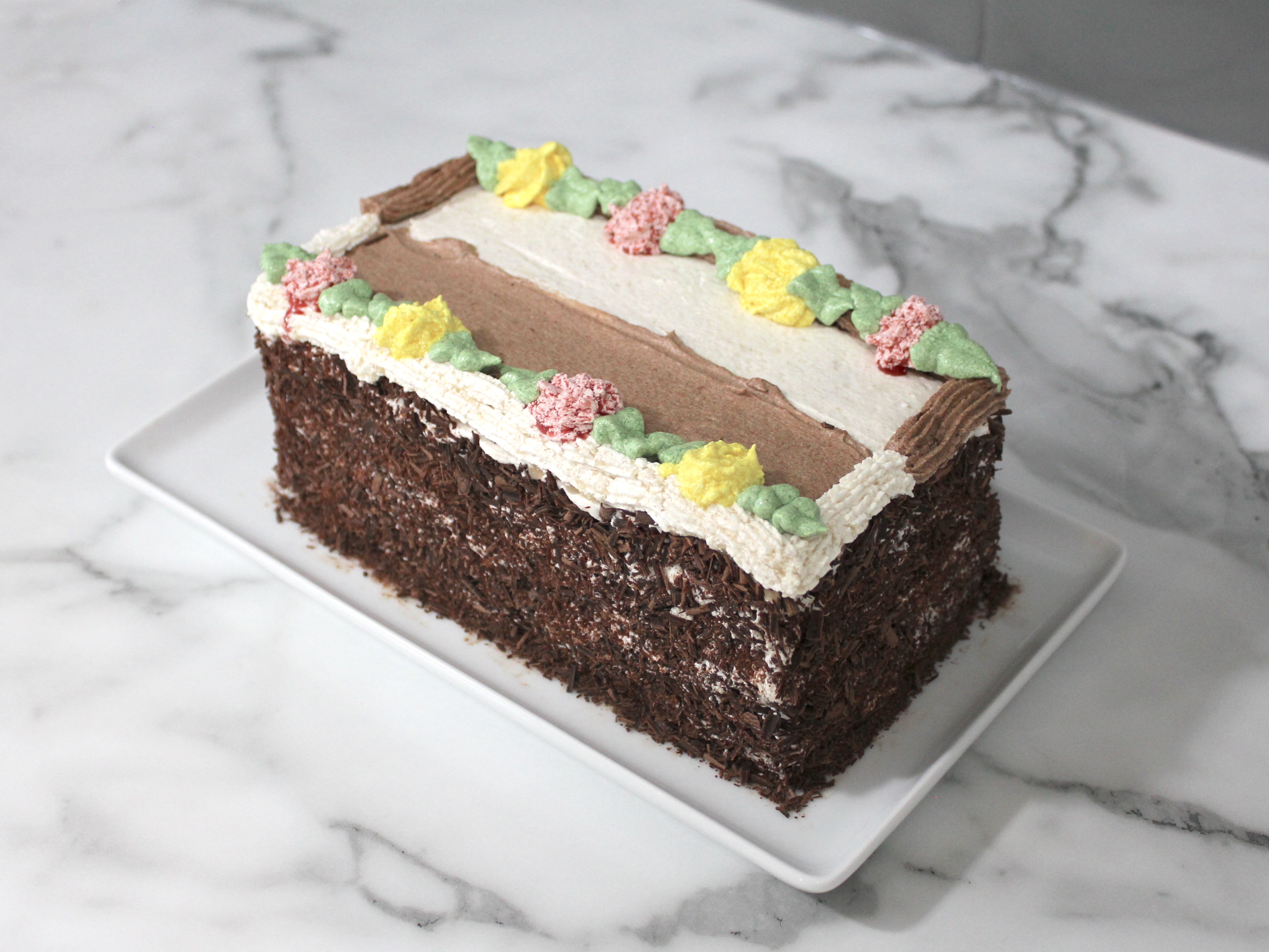 Full log cake. A 9-inch log cake covered in frosting and chocolate shavings. Top of cake has 8 alternating frosting flowers, 4 yellow and 4 pink with green leaves.