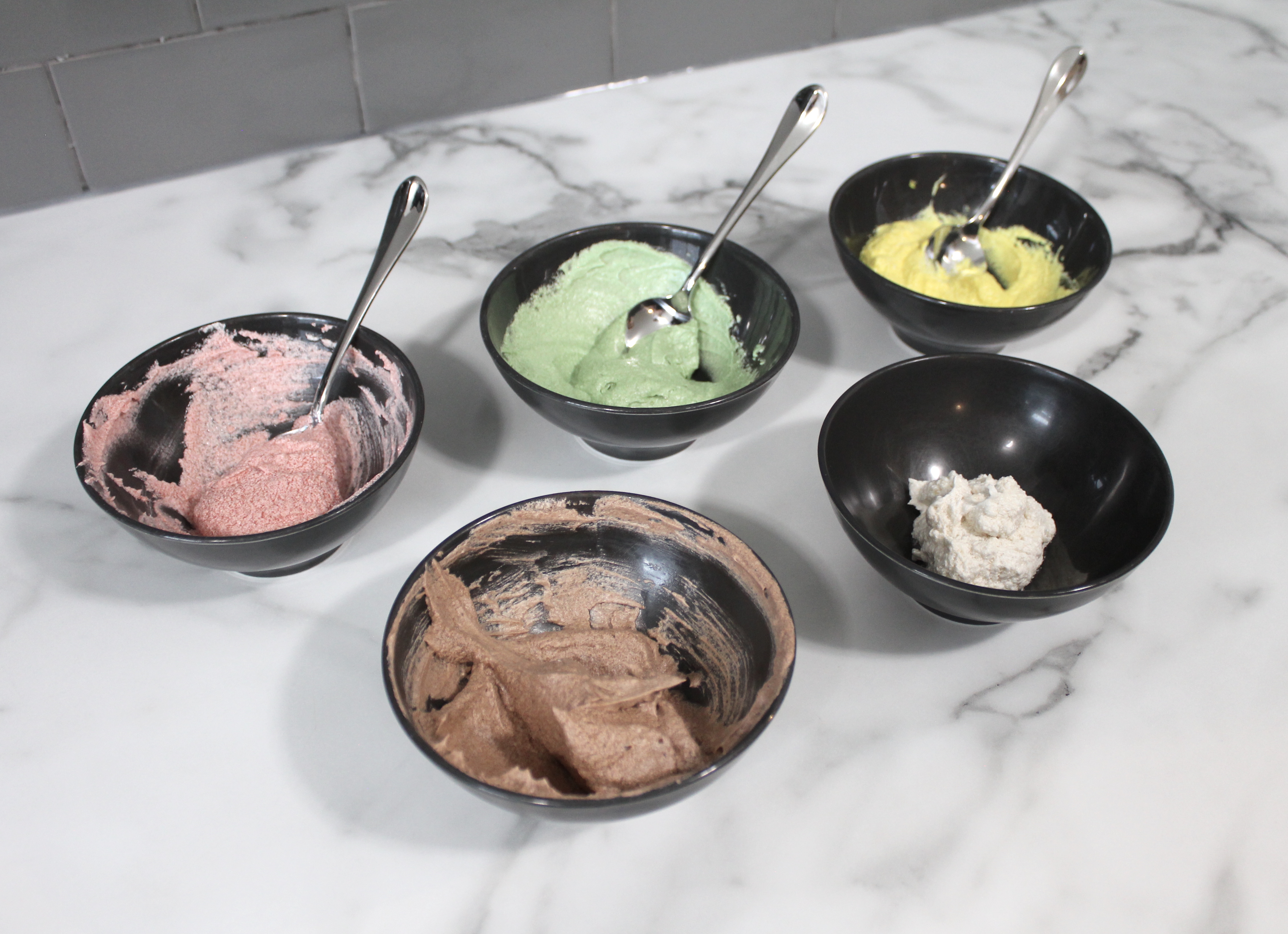 Five bowls with pink, green and yellow frosting, chocolate frosting and vanilla frosting.